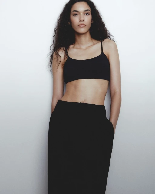 Fashion shot of a slim adult woman with curly dark hair wearing a black crop top and midi skirt against a white background.