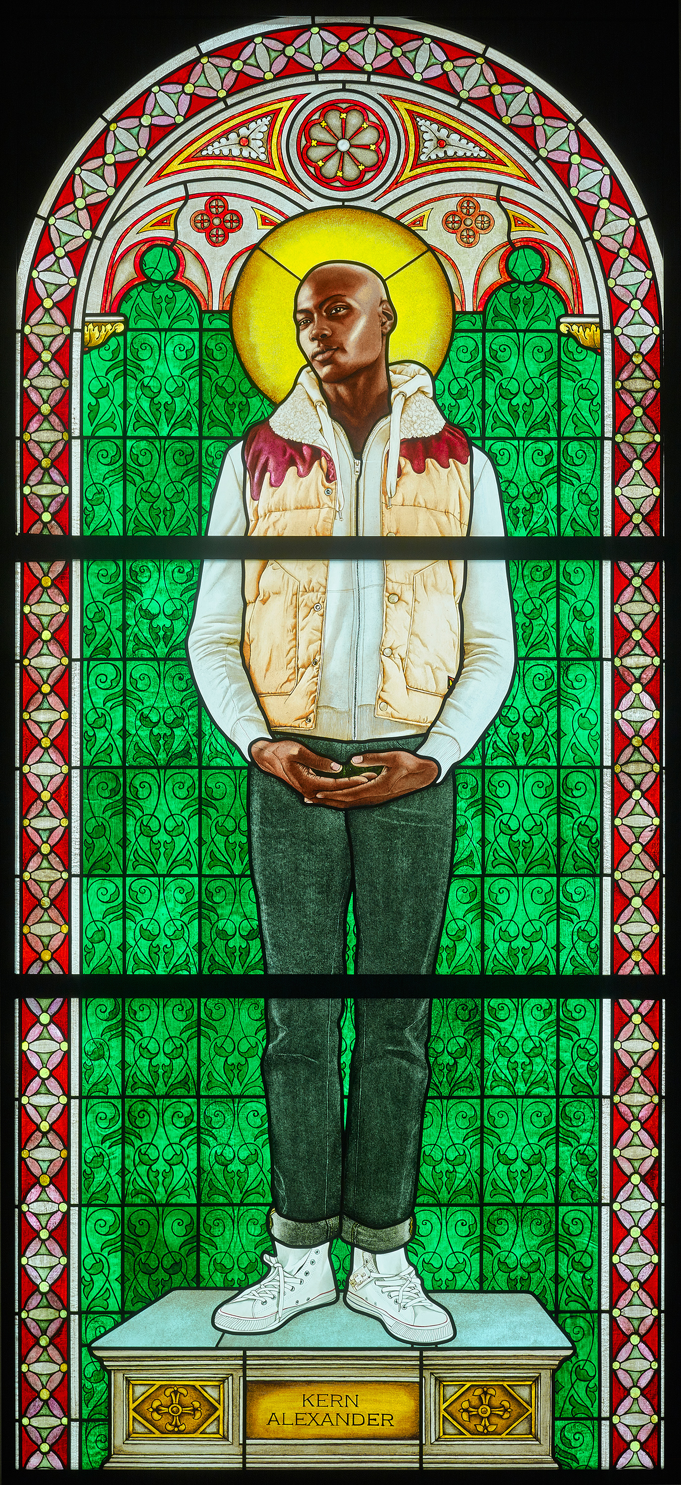 Stained glass artwork of a Black man with a halo wearing casual clothes, standing on a pedestal inside an architectural structure.