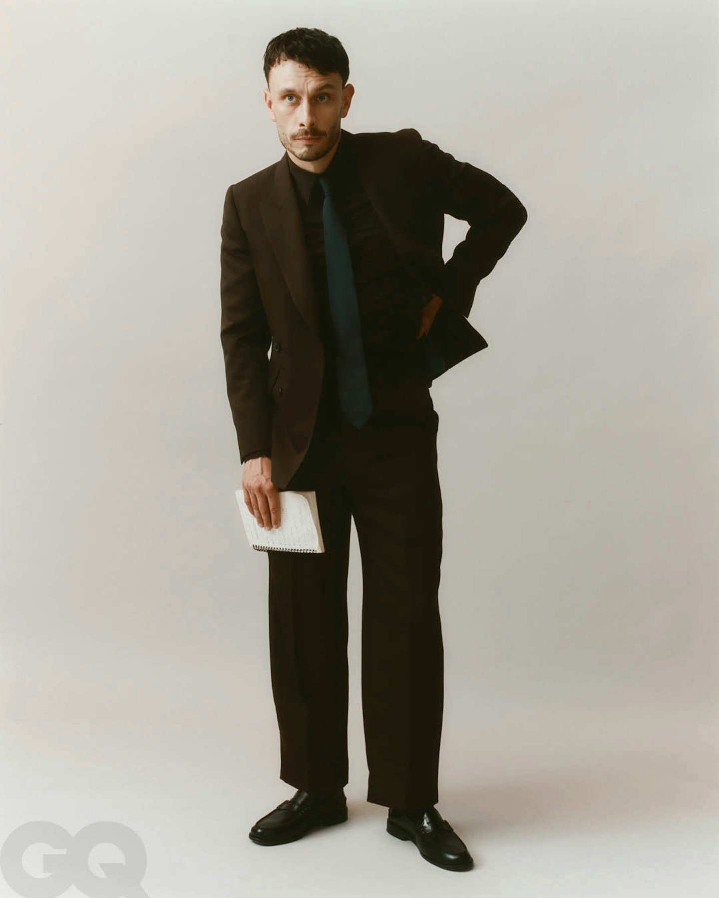 Adult man in dark suit, shirt and tie poses in studio with GQ logo visible at the bottom left in full-length shot.