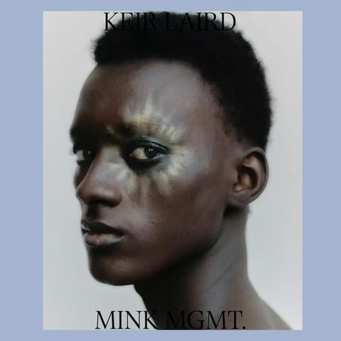 Close-up portrait of a Black male fashion model with dramatic makeup, styled hair, and a light grey-blue background framing his face.