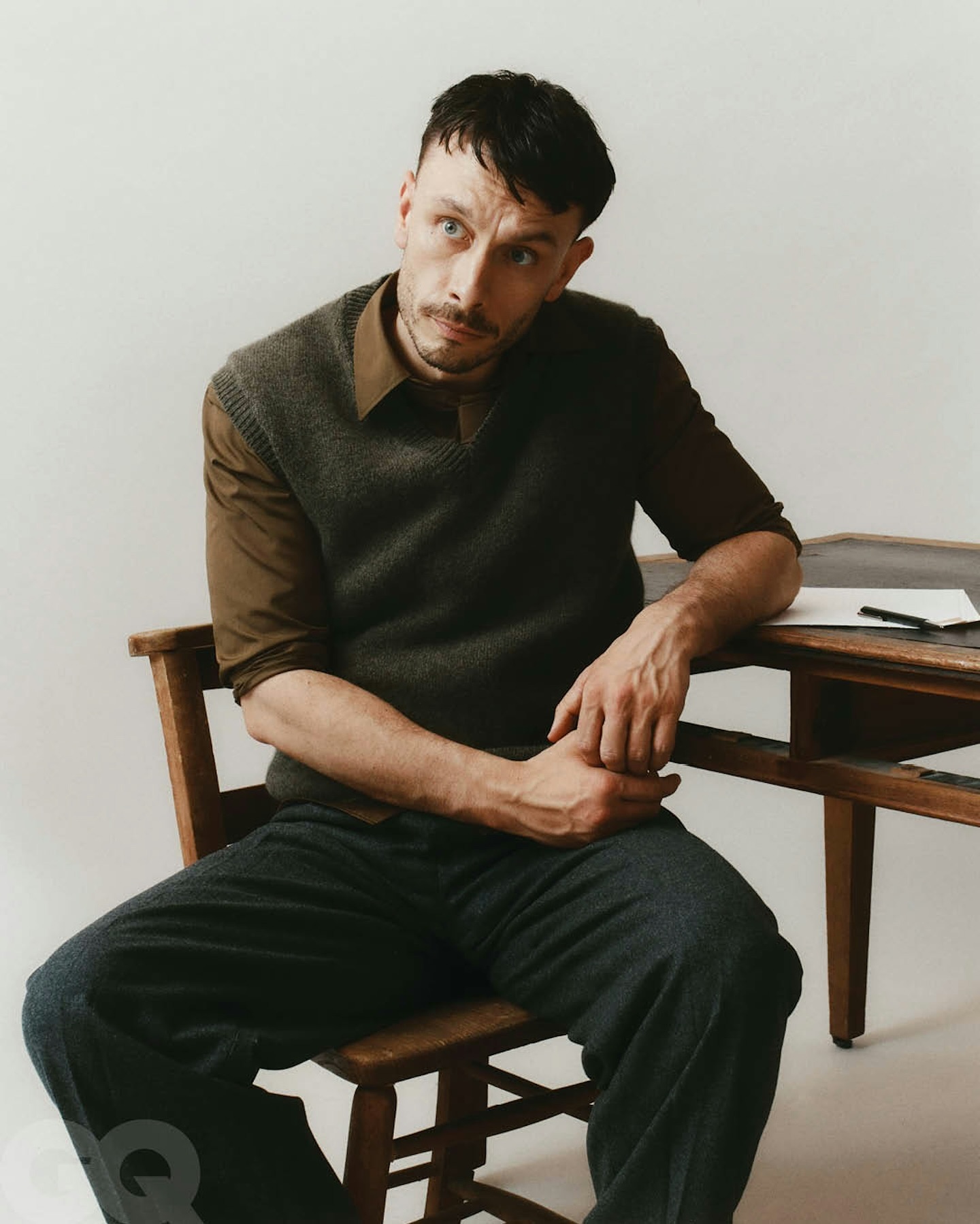Adult man sitting in a wood chair near a desk with paper and pen, wearing a collared shirt and sweater vest in a photo studio setting.