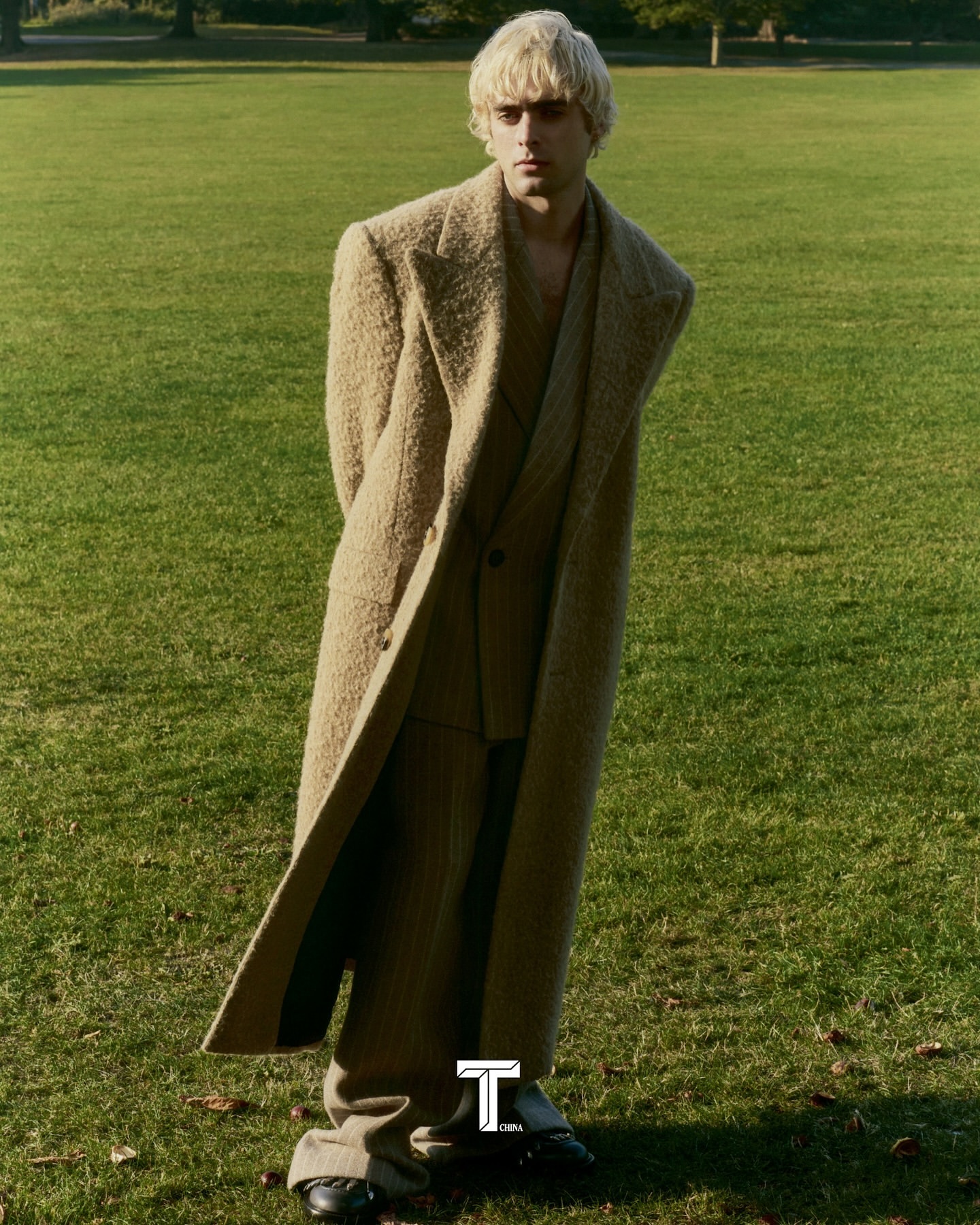 A fair-skinned male fashion model with blond hair poses in a wool coat and suit on grass in an outdoor setting with trees in the background.