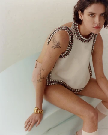 Woman wearing an embellished sleeveless top and gold cuff bracelet seated on a light green surface