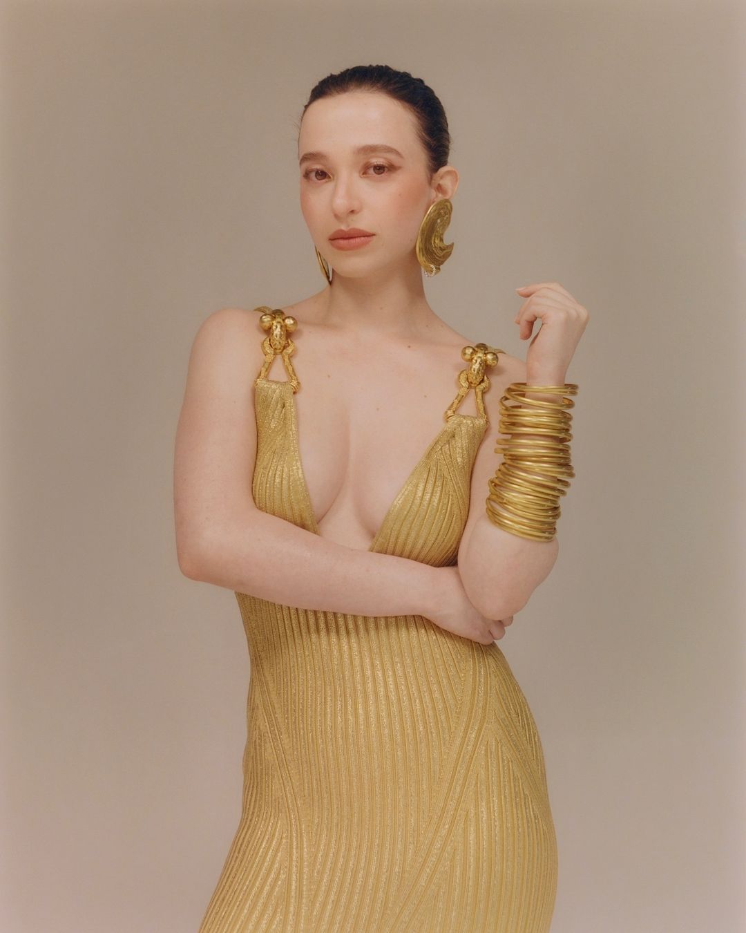 A woman wearing a gold dress and oversized gold jewelry poses in a photo studio with a light, neutral background for fashion editorial.