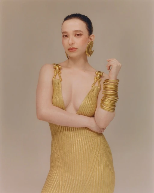 A woman wearing a gold dress and oversized gold jewelry poses in a photo studio with a light, neutral background for fashion editorial.