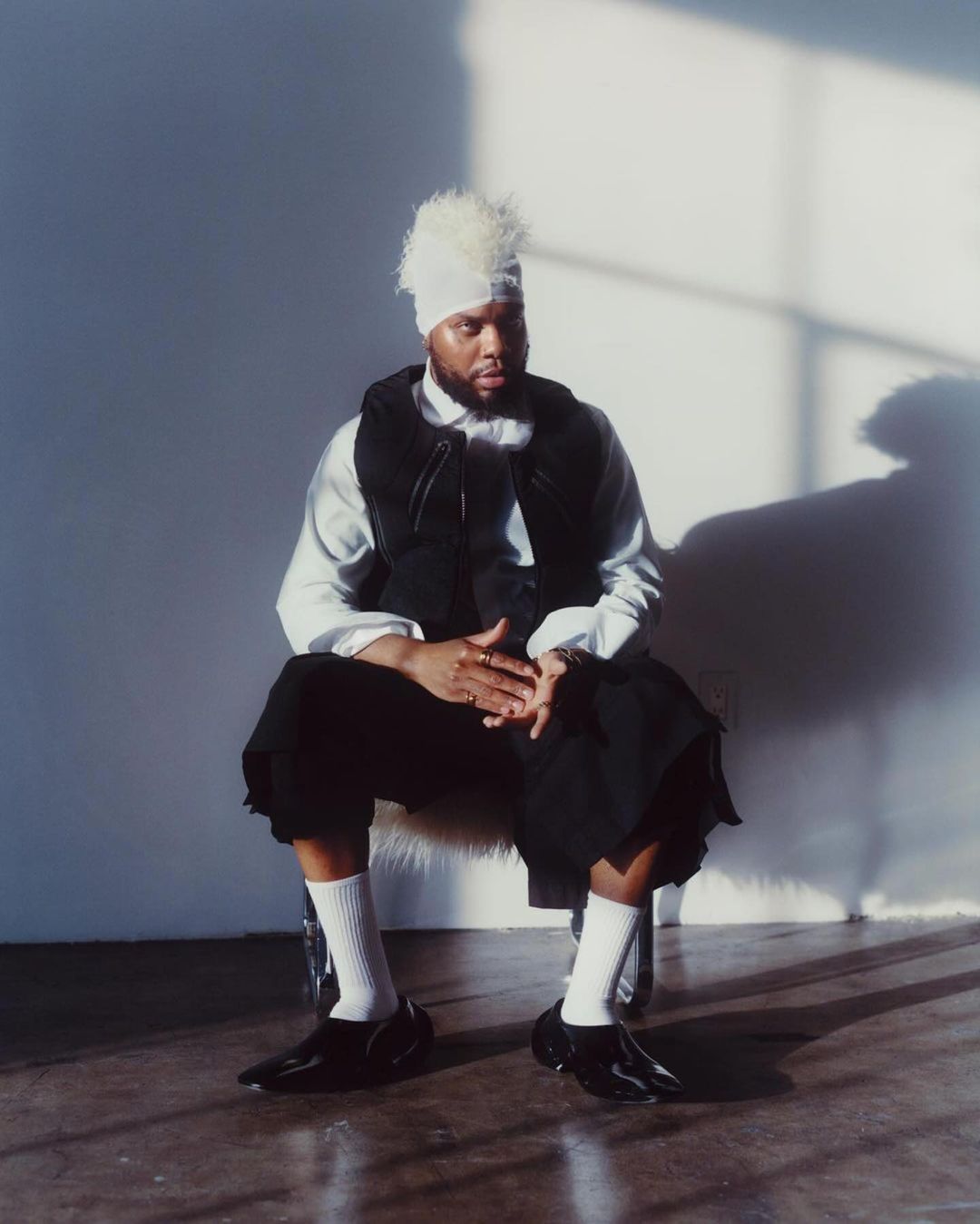 Black man seated wearing a white wrap headpiece, white shirt, black vest, black trousers, and white socks against a white wall.