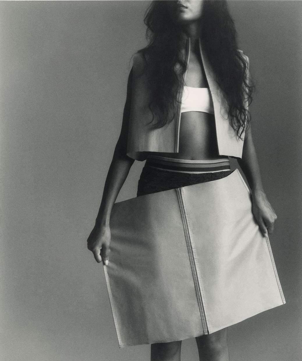A woman poses in a cropped sleeveless top and unique skirt against a grey backdrop, showcasing a minimalist and avant-garde fashion statement.
