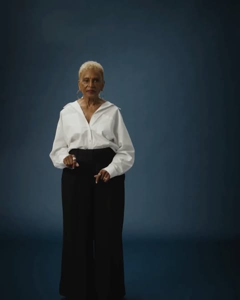 Older adult woman in a white button-down shirt and black trousers stands confidently in a studio portrait with dark blue background.