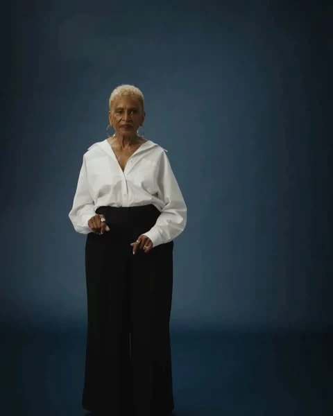 Older adult woman in a white button-down shirt and black trousers stands confidently in a studio portrait with dark blue background.