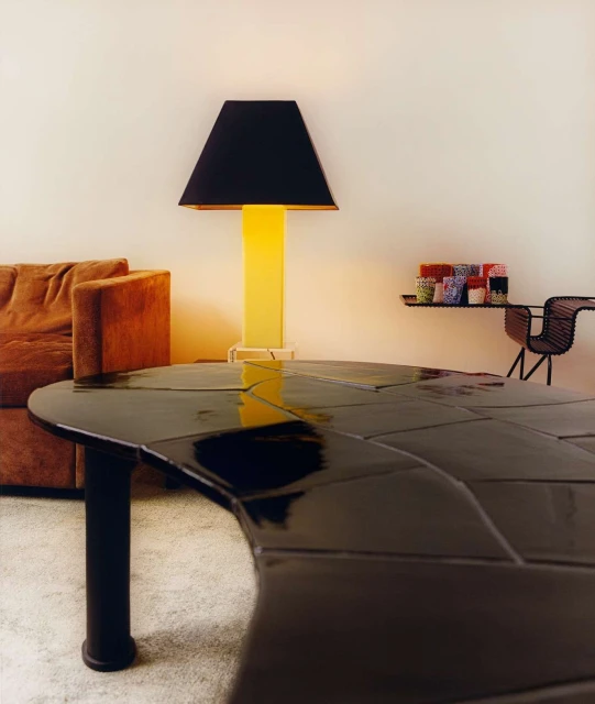 Interior with a black lacquered table, a yellow floor lamp with a black shade, and a brown sofa against a neutral wall.