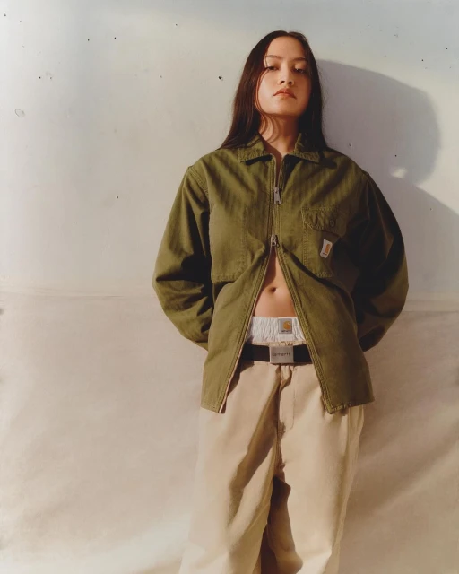 Woman with long hair in olive jacket and khaki pants stands in front of a mottled off-white wall with shadows. Fashion portrait.