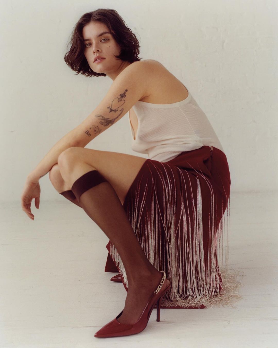 Woman in a studio setting wearing a burgundy skirt with white fringe, top, knee socks, and heels while crouching in the foreground.