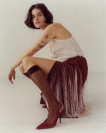 Woman in a studio setting wearing a burgundy skirt with white fringe, top, knee socks, and heels while crouching in the foreground.
