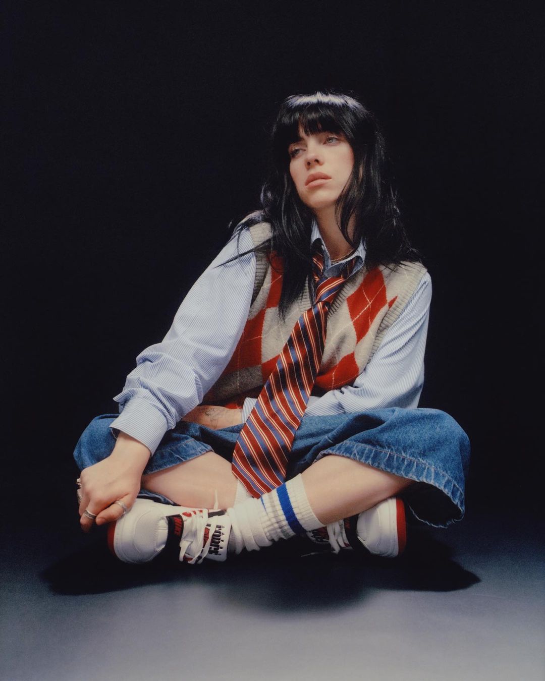 Portrait of a woman sitting cross-legged in a studio wearing a patterned vest, striped tie, baggy jeans, and socks with sneakers on a dark background.