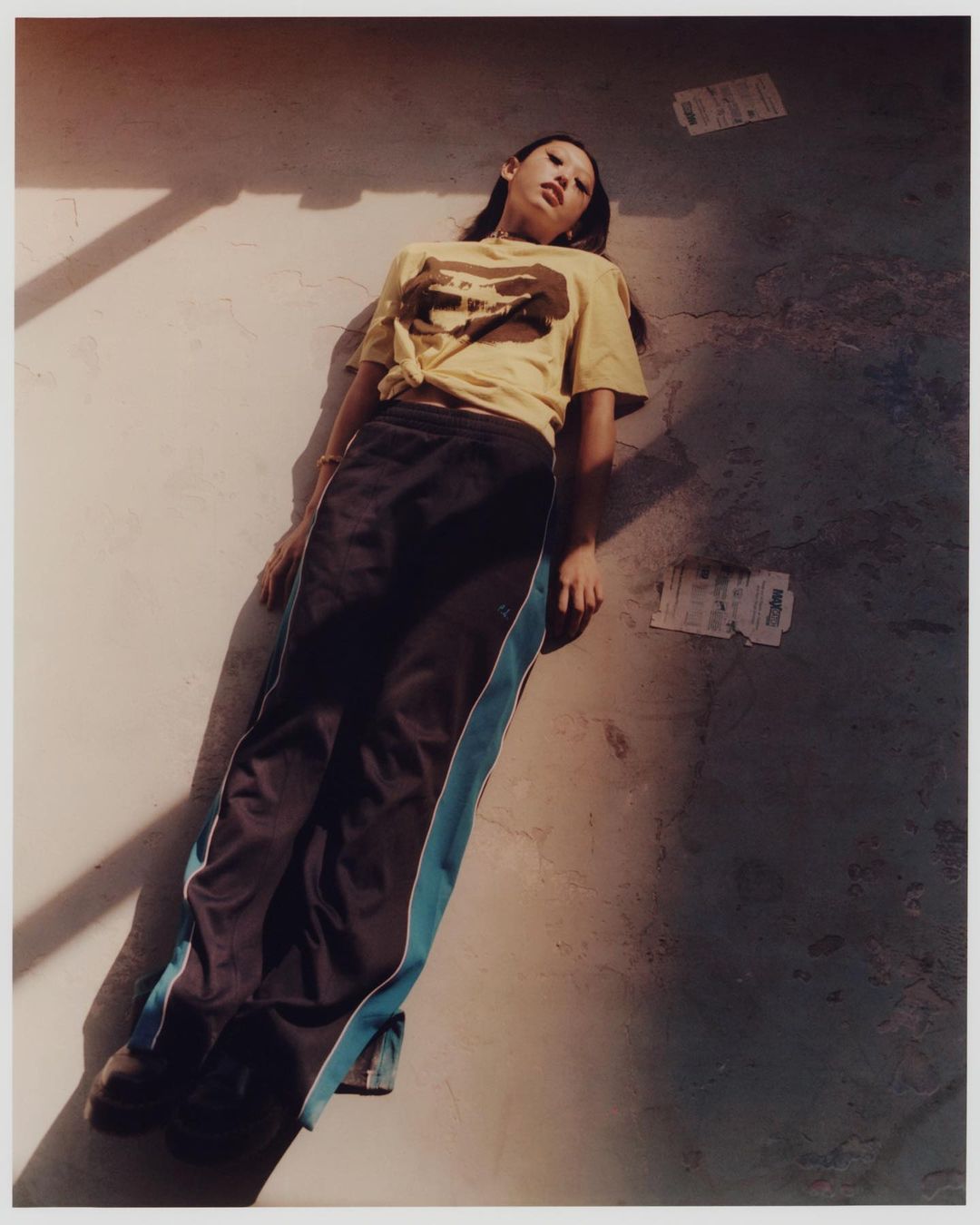 Asian woman with long dark hair, wearing a yellow graphic tee and black track pants, lies on a concrete surface in outdoor setting.