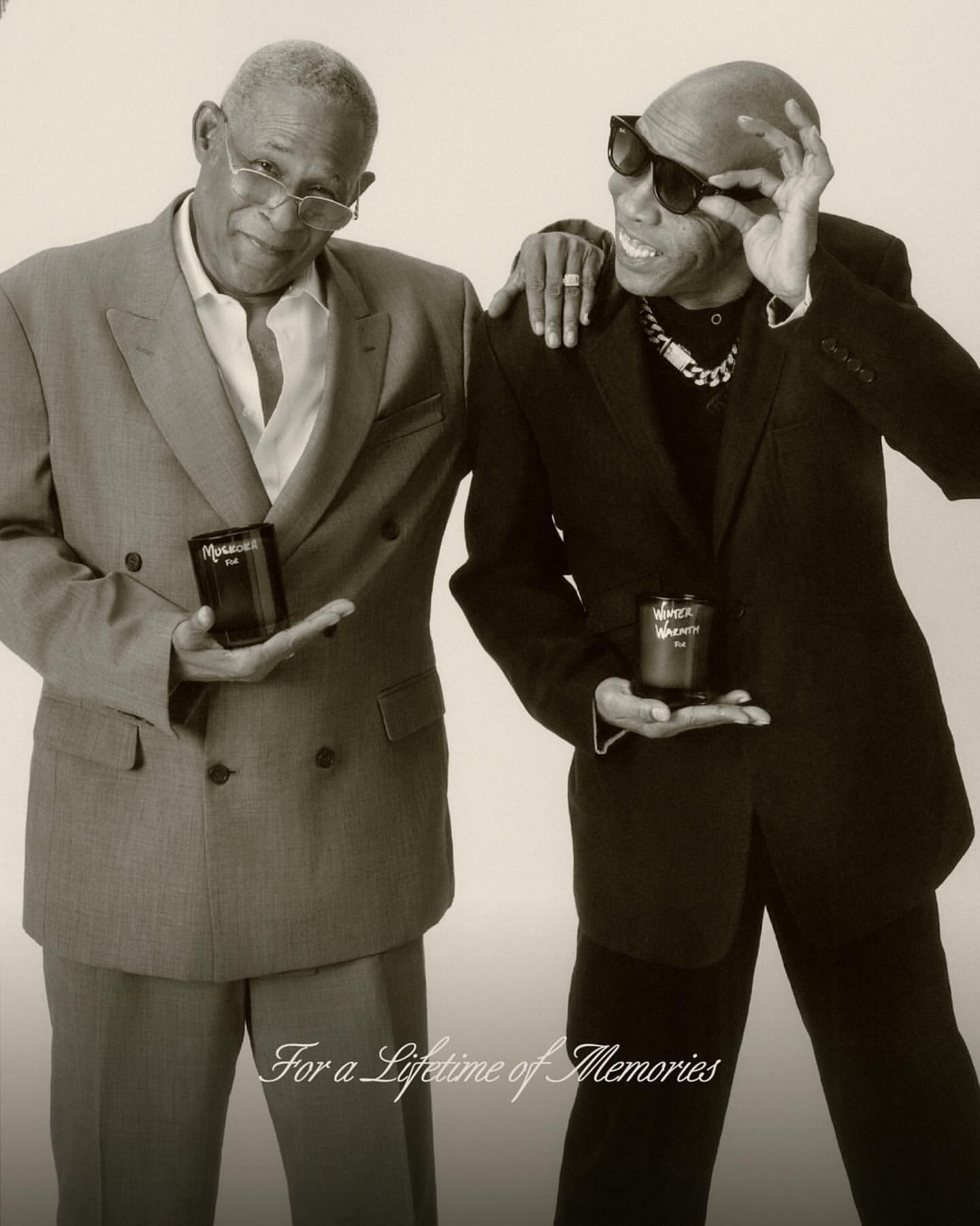 Two stylish adult males pose indoors, each holding a product, wearing suits and sunglasses with a light-colored backdrop.
