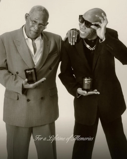 Two stylish adult males pose indoors, each holding a product, wearing suits and sunglasses with a light-colored backdrop. 