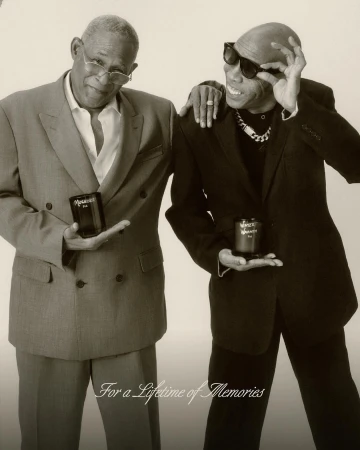 Two stylish adult males pose indoors, each holding a product, wearing suits and sunglasses with a light-colored backdrop. 