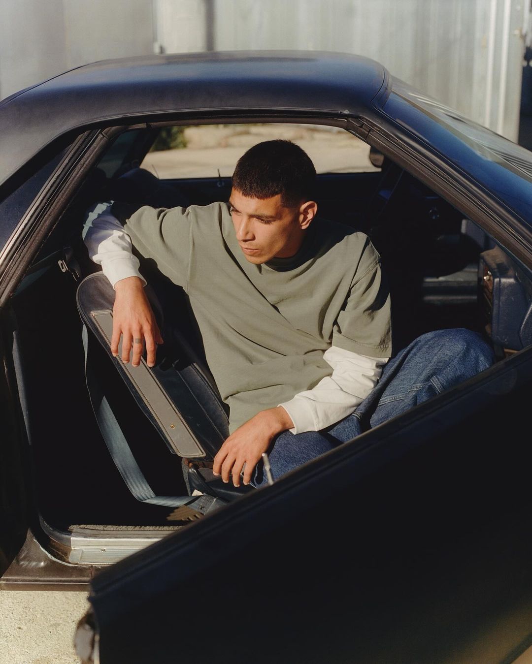 Adult male sits in a vintage car with an open door on a sunny day, wearing a green tee shirt and jeans in an outdoor setting.