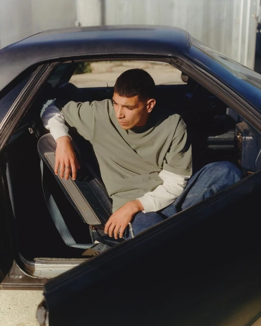 Adult male sits in a vintage car with an open door on a sunny day, wearing a green tee shirt and jeans in an outdoor setting.