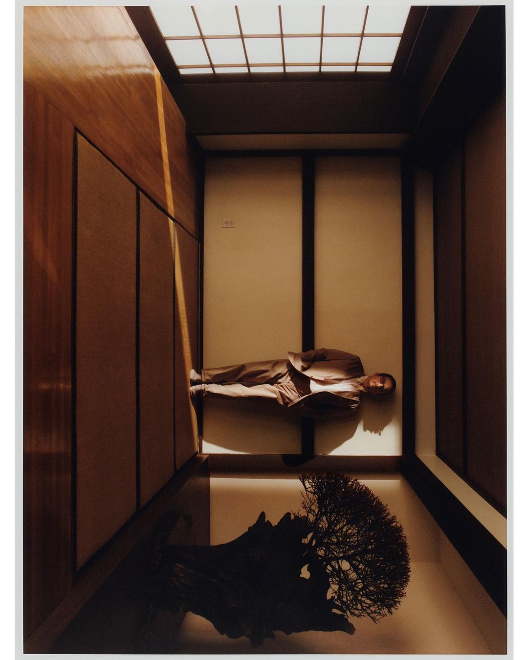 Adult male in suit lying indoors in a modern architectural setting, featuring wood panels, light, bonsai tree, and minimalist design.