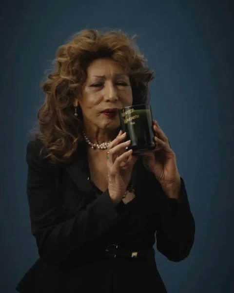 Elegant mature woman in black blazer holding a green candle, indoor against a blue background in a beauty and lifestyle shot.