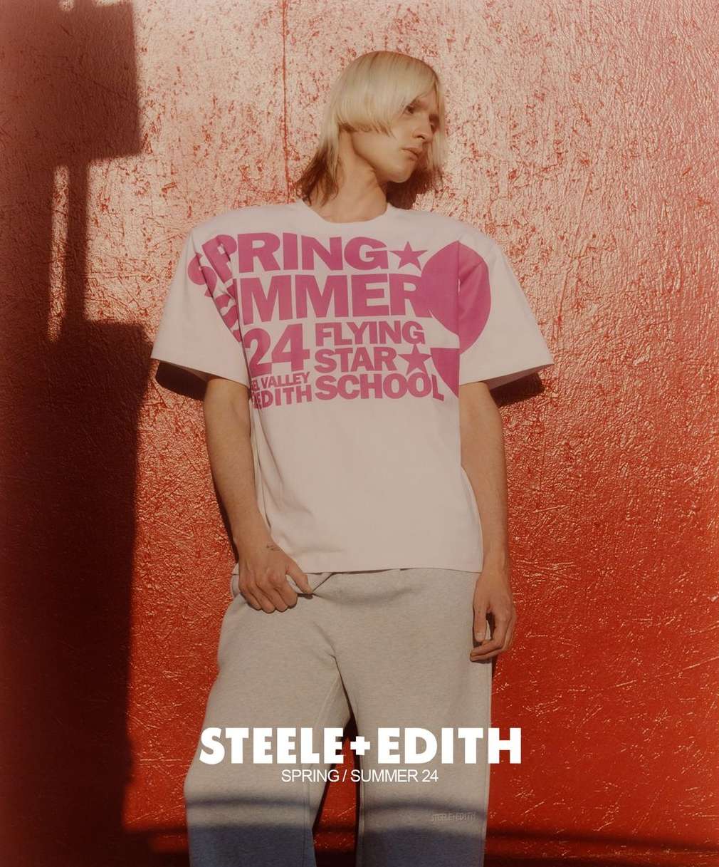 Young adult fashion model wears a white graphic tee and grey sweatpants, posing against a textured orange wall in soft lighting.