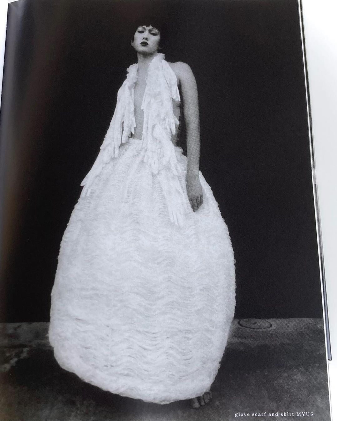 A person posing in an avant-garde dress featuring white gloves, against a dark background in a fashion editorial shot.