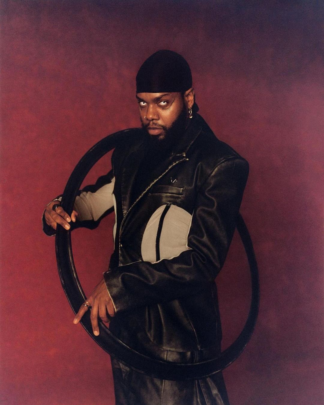 Stylish portrait of a black male in a leather jacket holding a circular object against a red background in a studio setting.