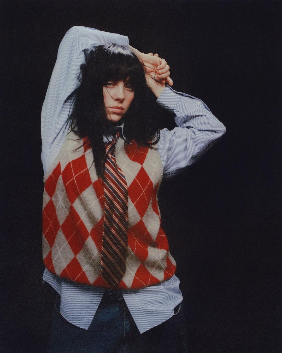 A woman with dark hair poses in a blue striped shirt, red argyle sweater vest, and tie against a dark backdrop in a studio.