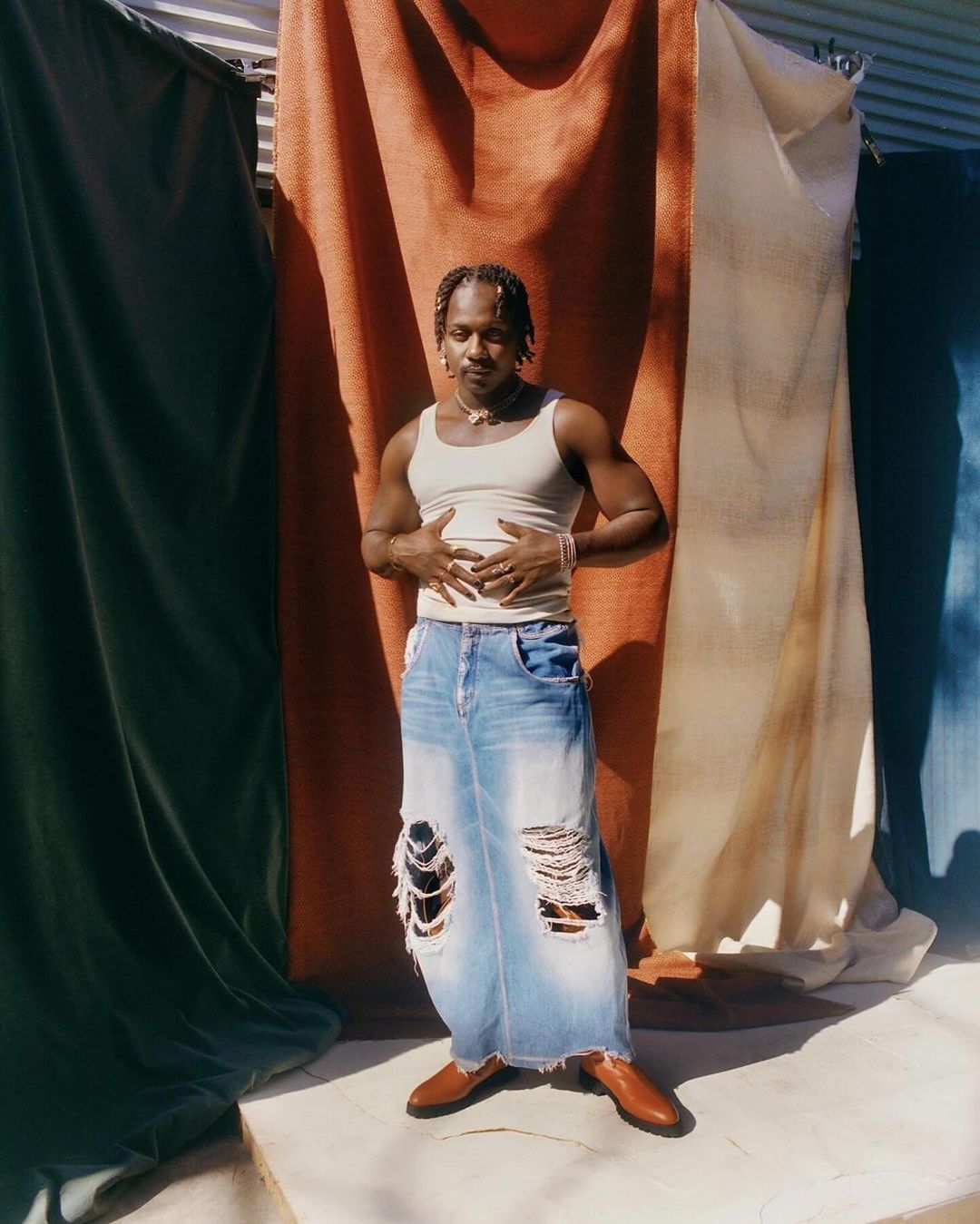 Man stands in tattered long denim skirt with tan tank top and shoes against draped backdrop in full shot outdoors during the day.