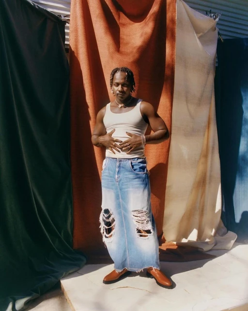 Man stands in tattered long denim skirt with tan tank top and shoes against draped backdrop in full shot outdoors during the day.
