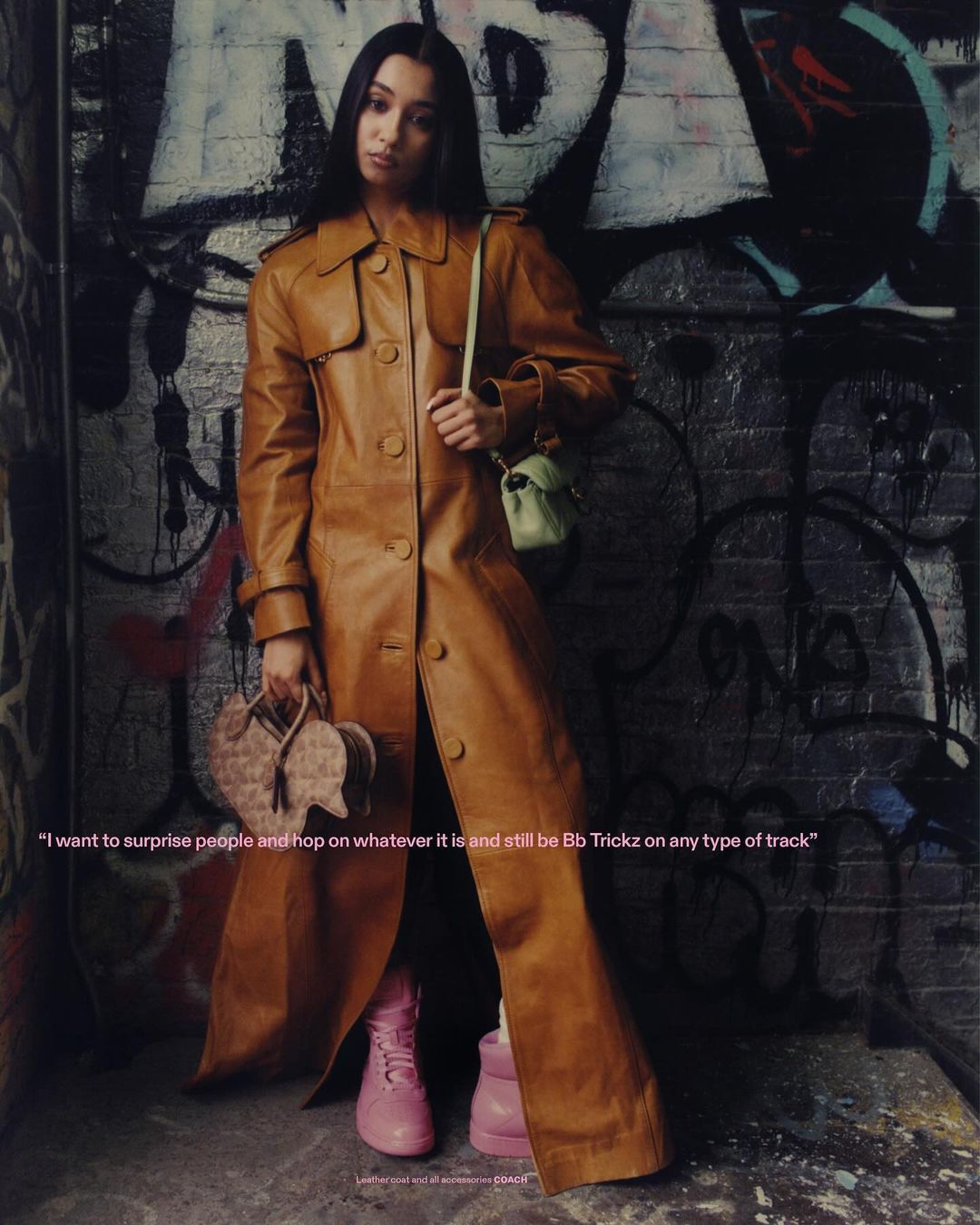 A woman in a tan trench coat poses against a graffiti wall, holding heart-shaped purse and wearing pink boots in this urban fashion shot.