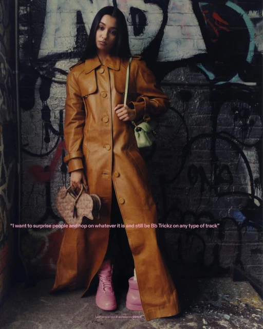 A woman in a tan trench coat poses against a graffiti wall, holding heart-shaped purse and wearing pink boots in this urban fashion shot.