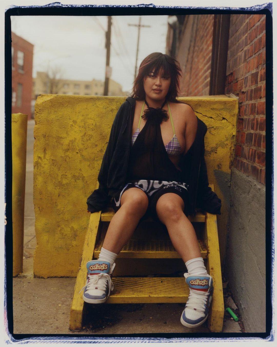 Young woman sitting outdoors on yellow metal steps in casual fashion with athletic shoes, shorts, and a black cover-up.