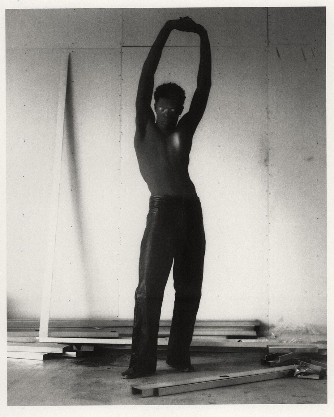 Striking black and white shot of a muscular adult male with arms raised, posing shirtless in a sparsely decorated interior.