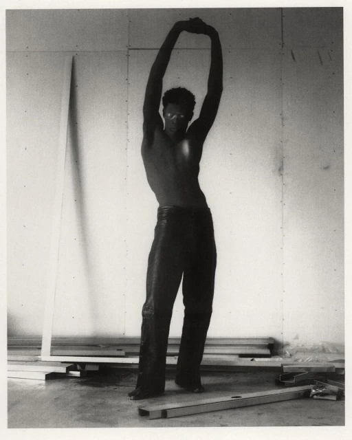 Striking black and white shot of a muscular adult male with arms raised, posing shirtless in a sparsely decorated interior.