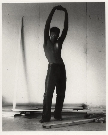 Striking black and white shot of a muscular adult male with arms raised, posing shirtless in a sparsely decorated interior.
