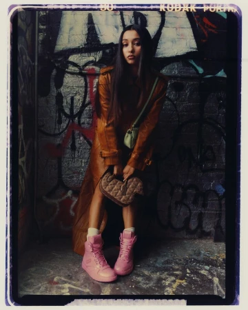 A fashionable young woman poses in front of graffiti art wearing a tan coat, pink sneakers, and carrying a small quilted purse.