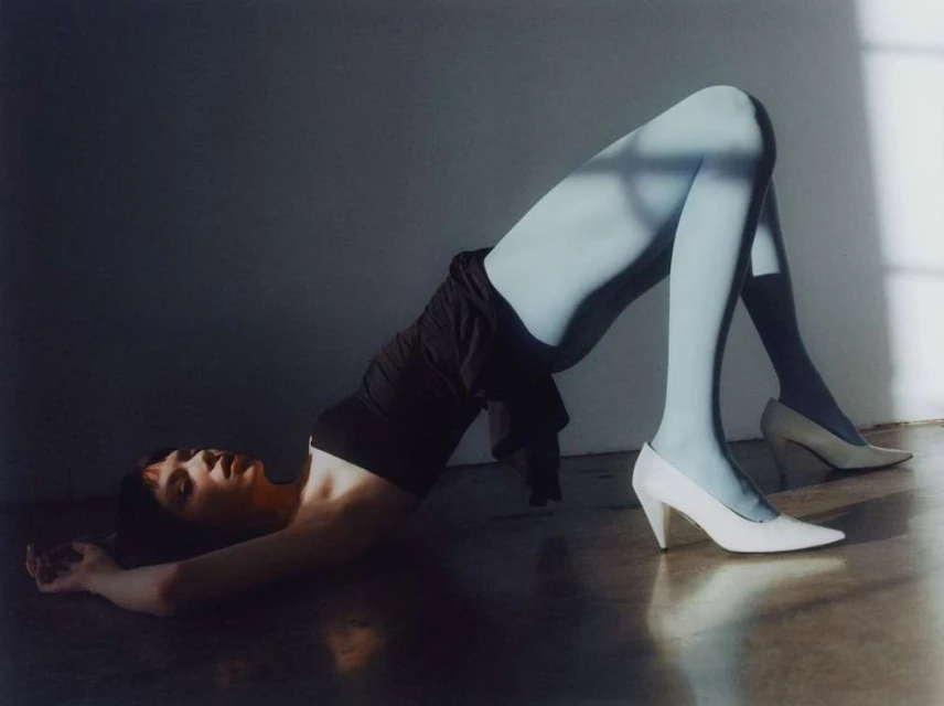 Woman in blue tights and white heels poses on floor with light and shadow play, wearing black top in a fashion shot.