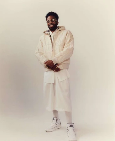 Portrait of a Black man wearing a white puffer jacket and shorts standing against a plain backdrop