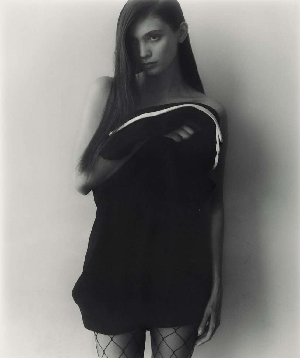 Monochrome portrait of a woman with long dark hair wearing fishnet tights and holding a dark garment against a pale background