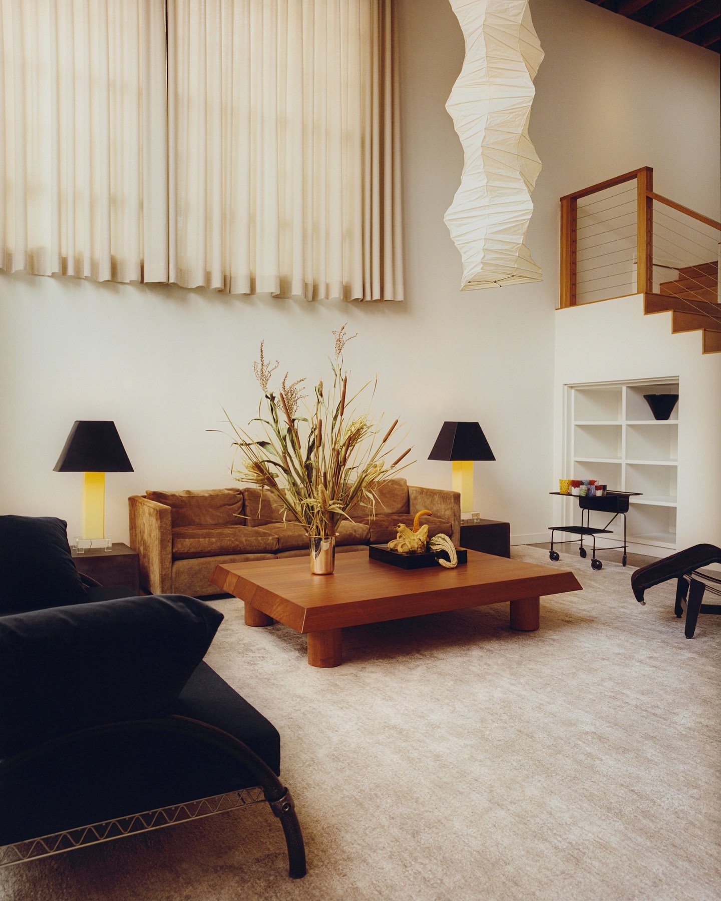 Living room interior featuring a brown sofa, wooden coffee table with vase, black chair, neutral carpet, and modern lighting fixtures.