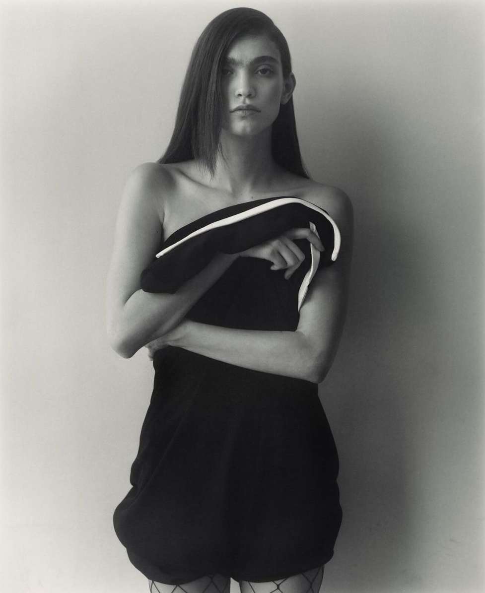 Monochrome studio portrait of a woman with long hair, posing in a short dark dress while holding a black and white garment.