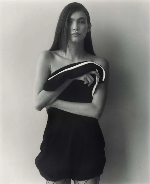 Monochrome studio portrait of a woman with long hair, posing in a short dark dress while holding a black and white garment.