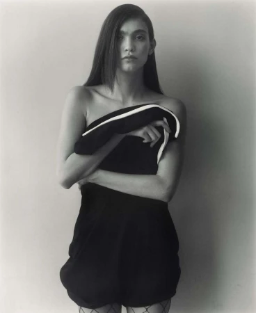 Monochrome studio portrait of a woman with long hair, posing in a short dark dress while holding a black and white garment.