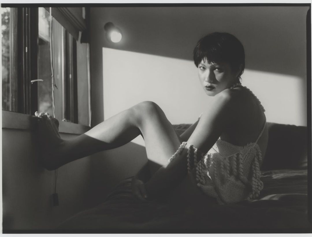 Monochromatic portrait of a short-haired woman in a crochet top sits on a bed by a window, illuminated by soft, directional lighting.
