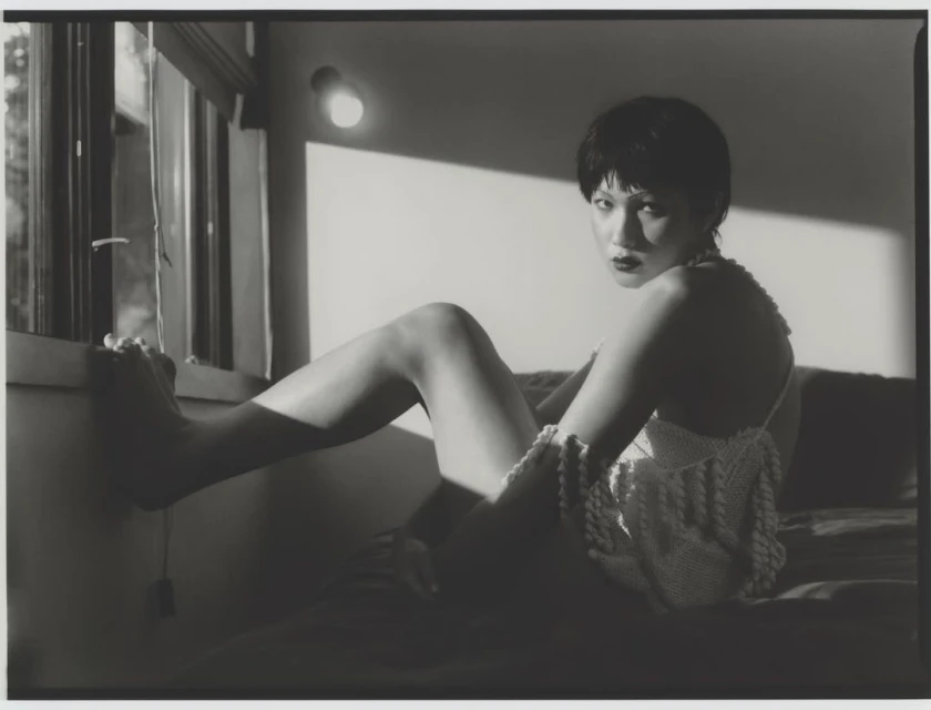 Monochromatic portrait of a short-haired woman in a crochet top sits on a bed by a window, illuminated by soft, directional lighting.
