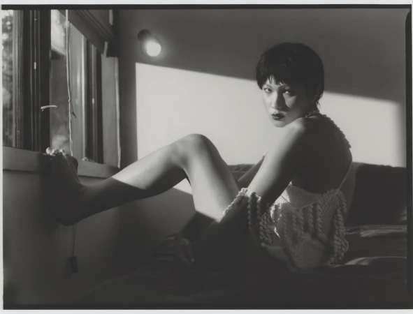 Monochromatic portrait of a short-haired woman in a crochet top sits on a bed by a window, illuminated by soft, directional lighting.