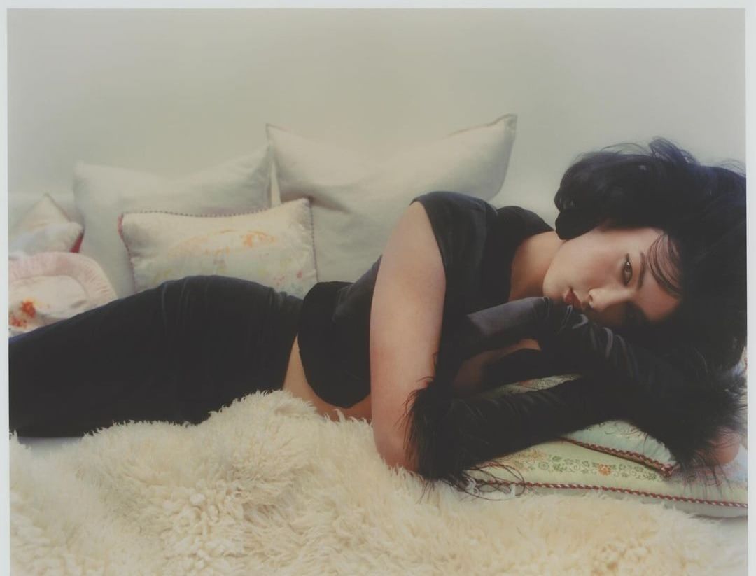 Woman with dark hair lies on a white fluffy rug, with decorative pillows behind her. She wears a black top with furry black arm coverings.